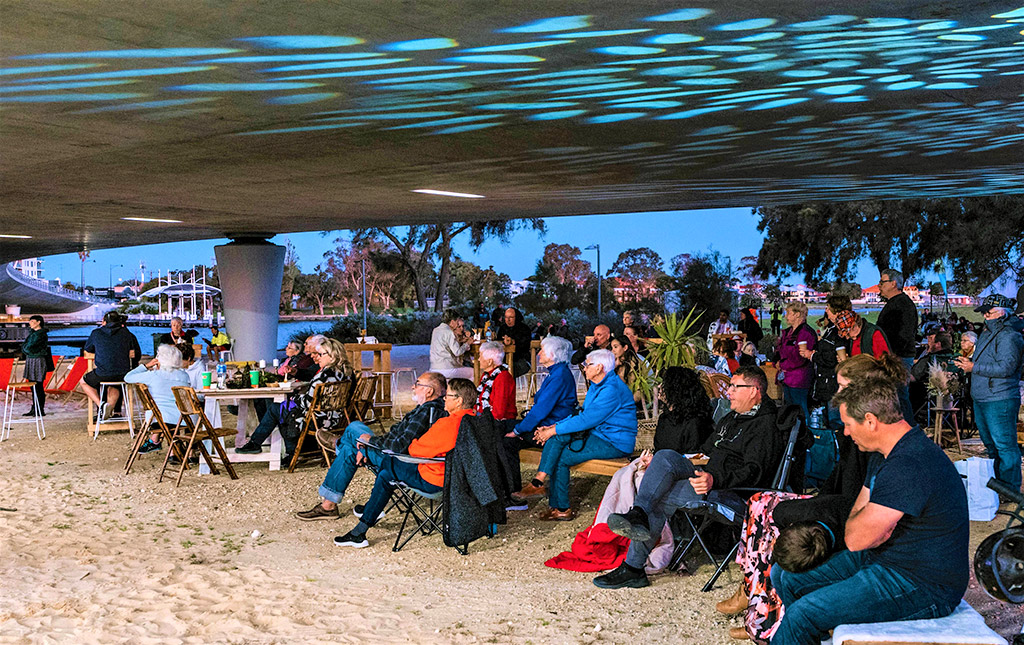 a crowd of people sit underneath a concrete bridge with theatrical stage lighting, watching a performance that is just out of frame