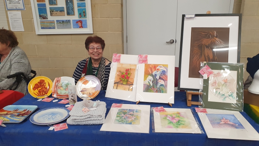 A woman poses with several painted artworks presented on a table with a blue tablecloth