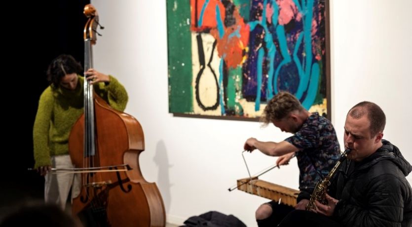 3 musicians play string and brass instruments together in front of a painting