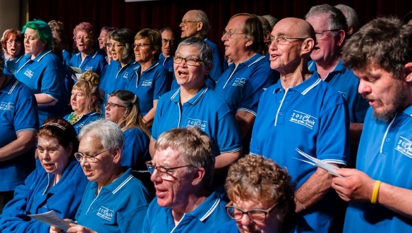 A group of older people in blue shirts stand in several lines, singing in a choir