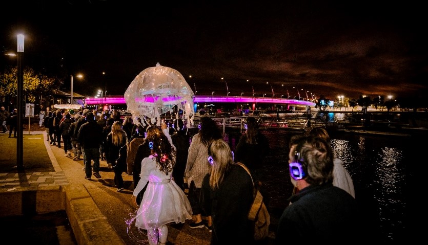 A group of people wearing headphones walk along a waterfront location with a bridge lit up with purple lights, led by a woman in a white dress with a white umbrella