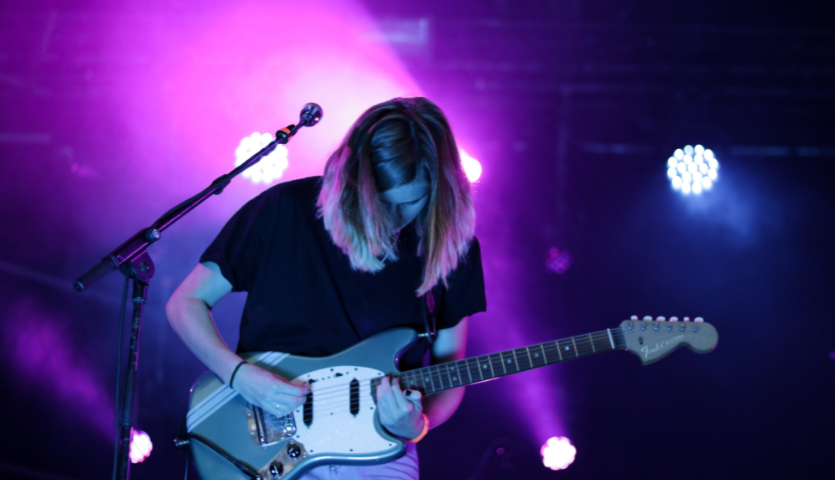 A young musician plays an electric guitar with their face downturned, a microphone in front of them and purple stage lighting in the background
