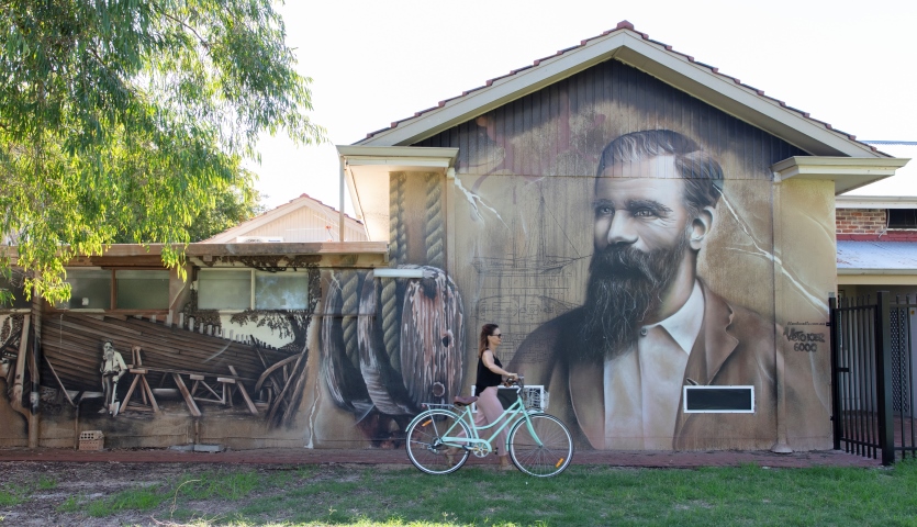 A woman on a bicycle rides past a mural of a bearded man on a building.