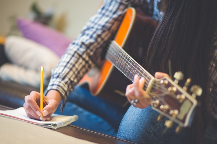 Person holding guitar in one hand and writing on paper with the other hand.