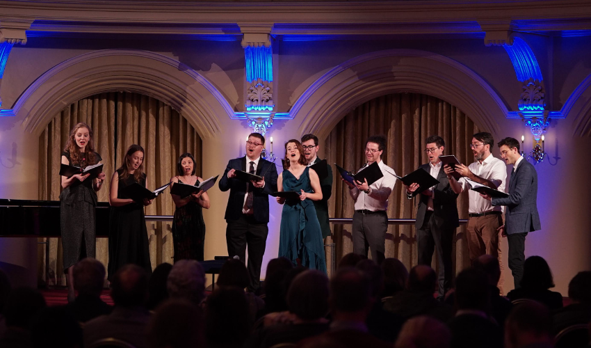 Overlooking the heads of an audience from behind with a choir performing on a lit stage in the background of the image