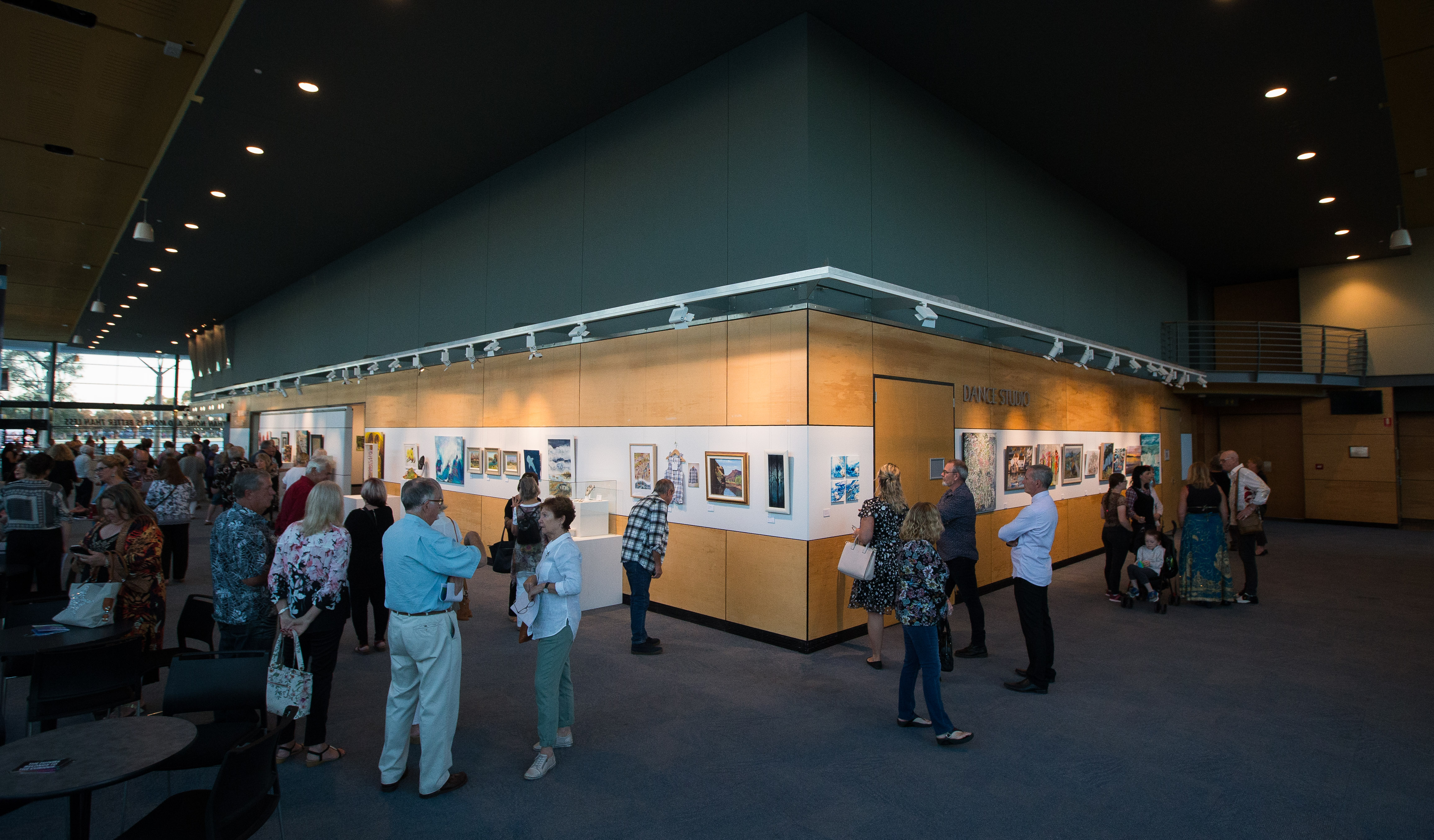 A wide angle photo of a gallery space at night, with lighting directed towards the artworks on the wall and many people standing around and admiring the artwork
