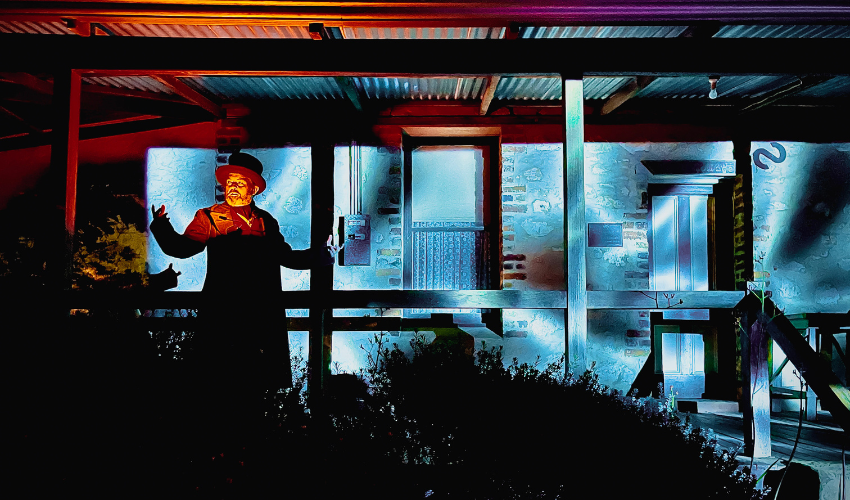 A man stands on a veranda with a cottage behind him that has been lit up with blue water like projections