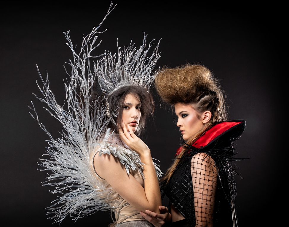 Two women in Wearable Art garments, one in a white dress and headpiece, the other in black and red garment.