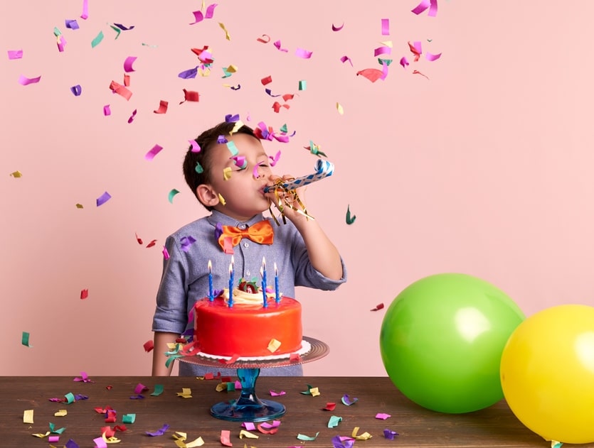 Male child celebrating a birthday with a cake, balloons, confetti and a party horn