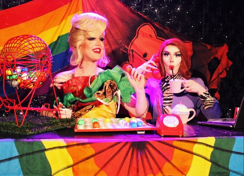 Two drag queens sitting at rainbow decorated table, one rolling bingo wheel and hosting the game, the other drinking a cup of coffee through a straw.