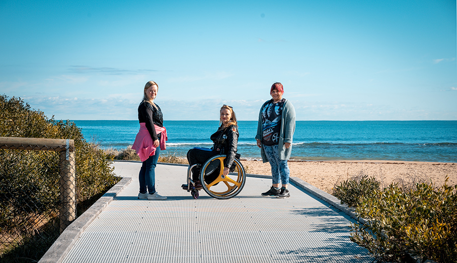 Three people on a boardwalk with the ocean in the background. One person is using a wheelchair.