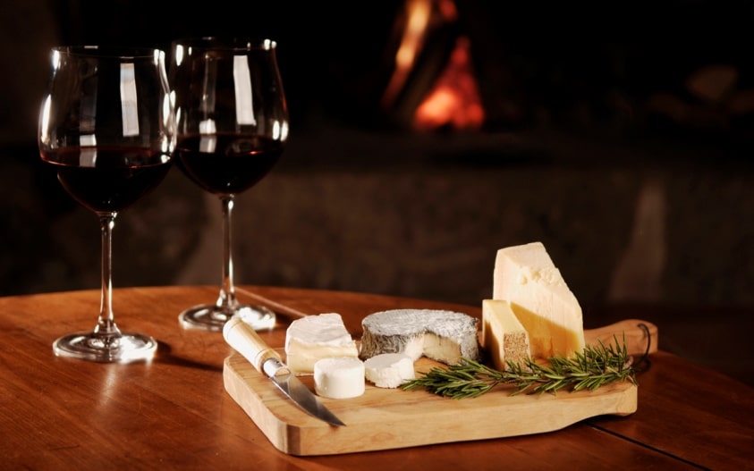 2 x glasses of red wine and a cheese platter sitting on a wooden table with a fire glowing in the background.