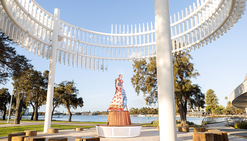 Model wearing colourful Wearable Art garment stands underneath Meeting Place public art on Mandurah foreshore