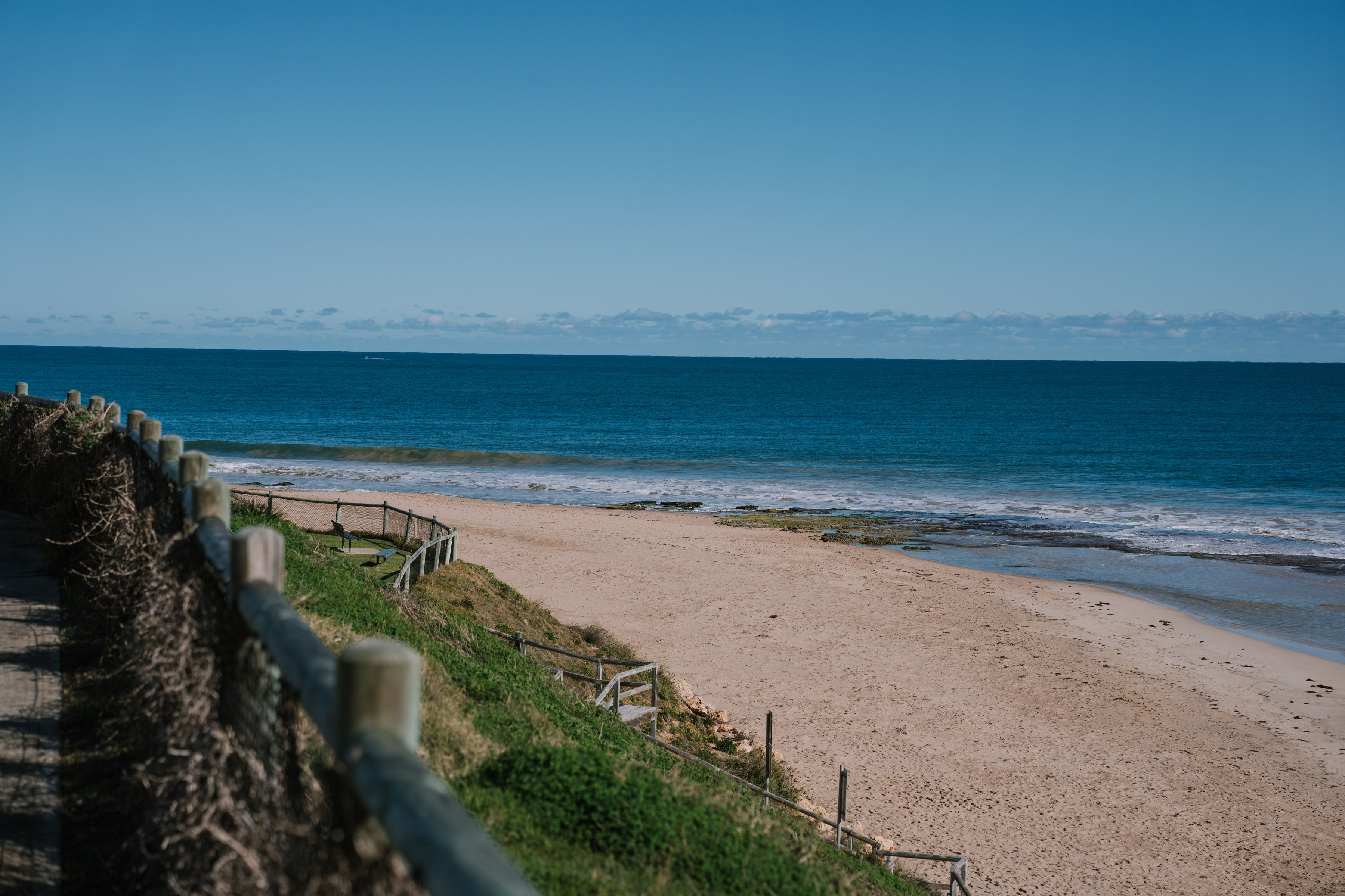 A sandy beach in the City of Mandurah with gentle waves meeting the shoreline under a clear blue sky. A wooden fence runs along a grassy path on the left side, overlooking the beach and ocean. The horizon is visible in the distance with a thin line of clouds.