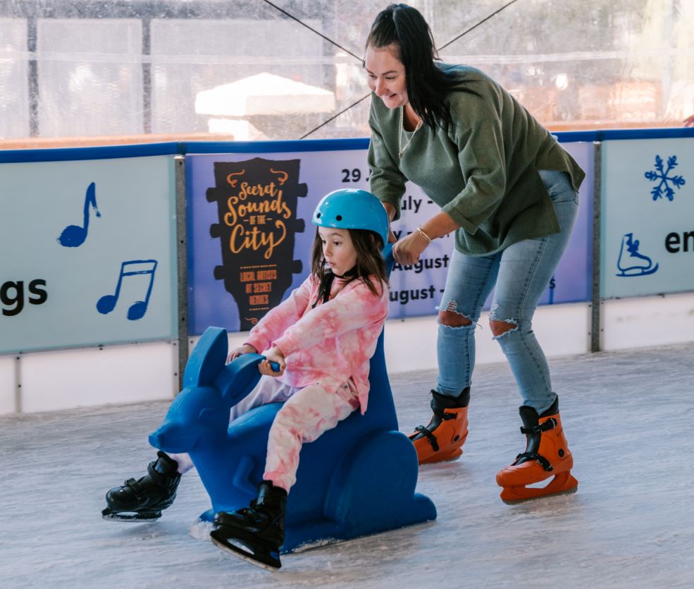 Child sits on skate aid while adult pushes from behind on ice rink