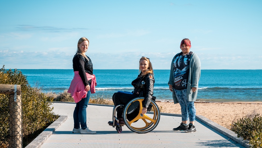 Three people on a beach access pathway with the beach in the background, one lady is in a wheelchair and the other two are standing.
