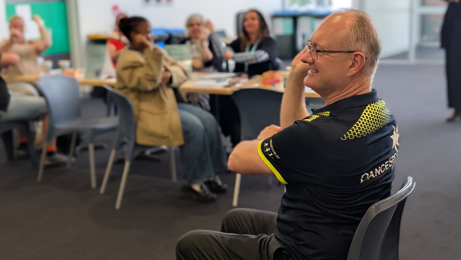 A smiling dance instructor sits on a chair facing a group of adults. The instructor wears a black and yellow Dancesport shirt and gestures with his arm while people watch and participate. The room has carpeted floors and large windows.