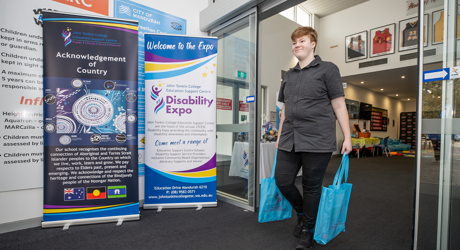 A person carrying two blue bags leaves the Disability Expo at Mandurah Aquatic and Recreation Centre. Event banners and display tables are visible.