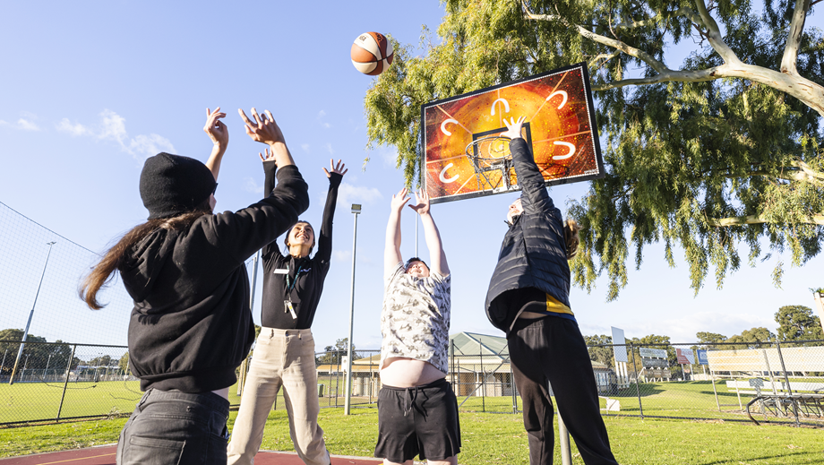 A group of Youth playing basketball, one shooting and three others jumping up to get the ball.