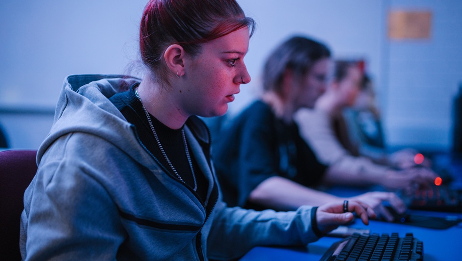 A teenage girl sitting at a computer typing on a keyboard with other teens sitting at computers beside her in the background.