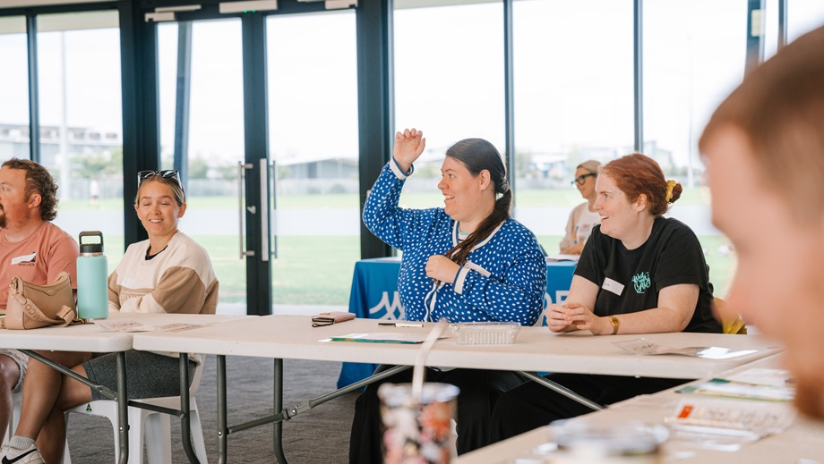 Adults sit at tables in a community room during a workshop. One person raises their hand while others listen.