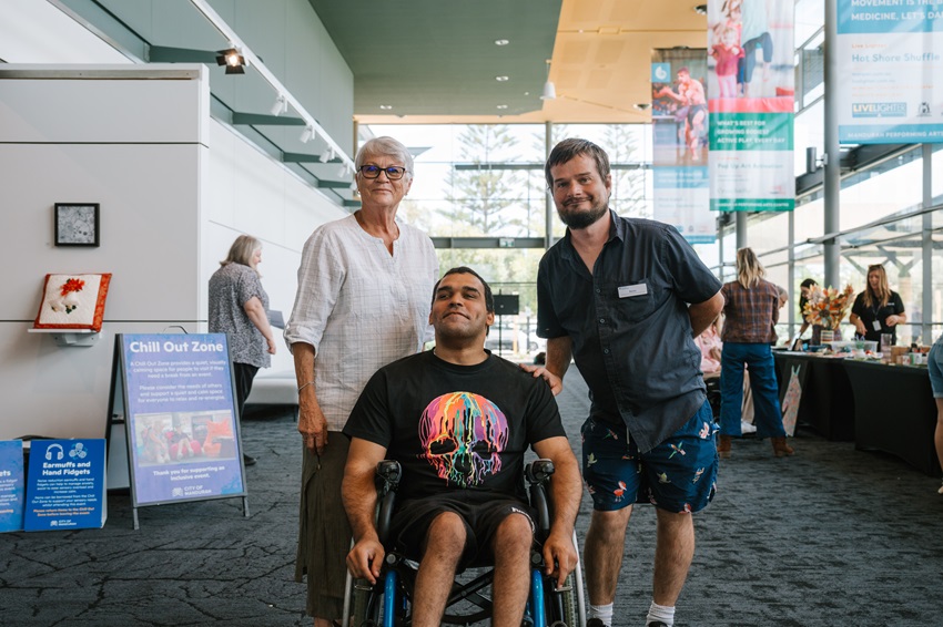 Three people smiling at the camera. One person is sitting in a wheelchair. In the background there are people and information stalls.
