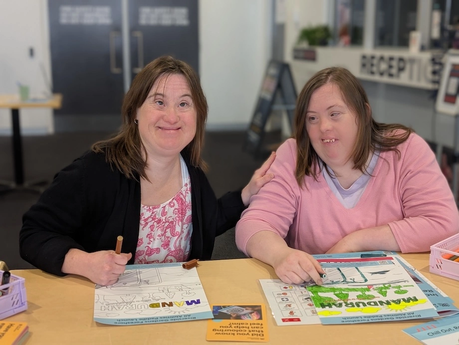 Two women sitting together at a table during a My Community Morning Tea session, using colouring sheets and pencils, with activity resources spread out in a community centre space