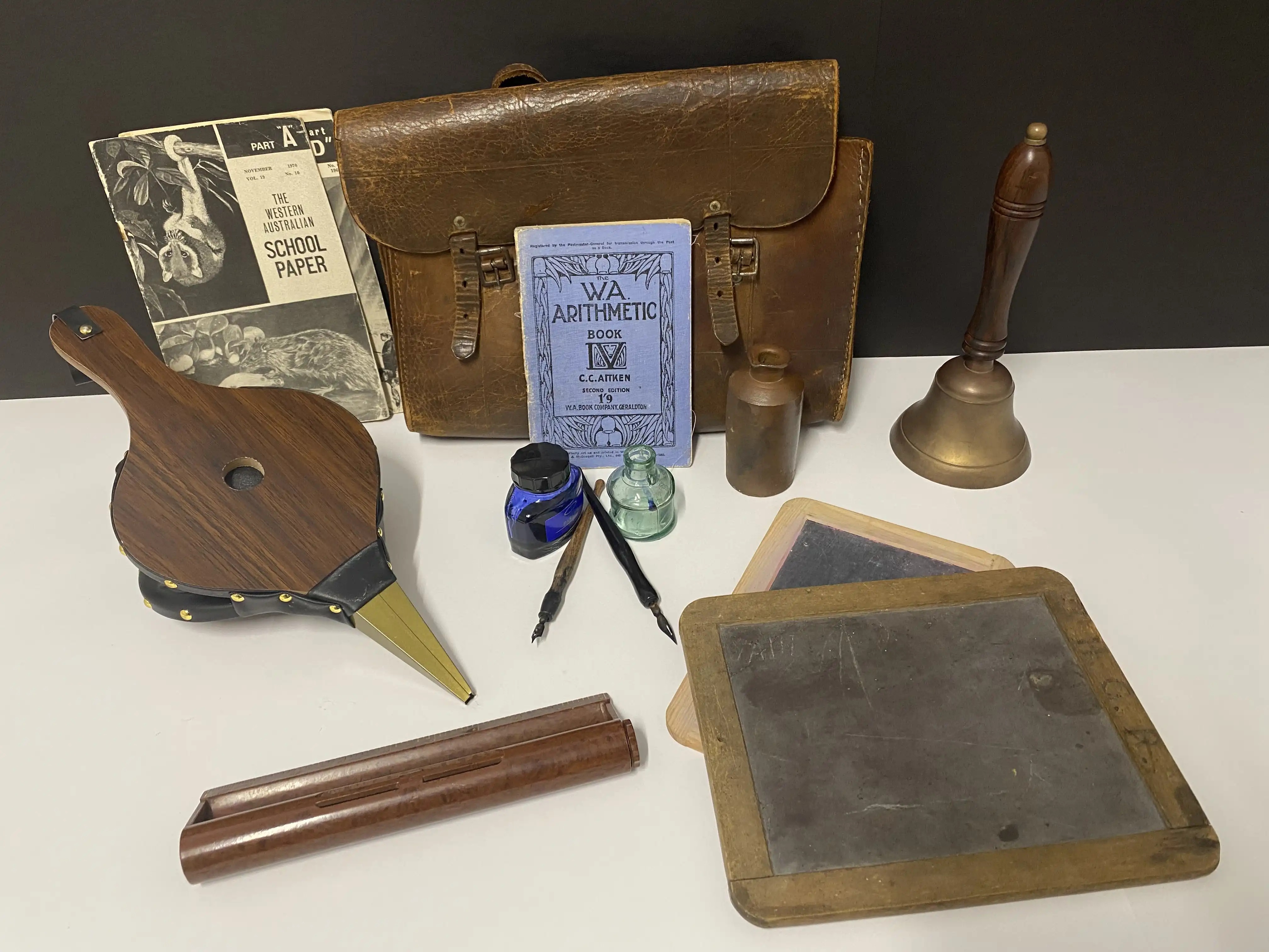 A collection of old-fashioned school items, including a leather satchel, slate boards, ink bottles with dip pens, a bell, a wooden bellows, and vintage exercise books arranged on a table